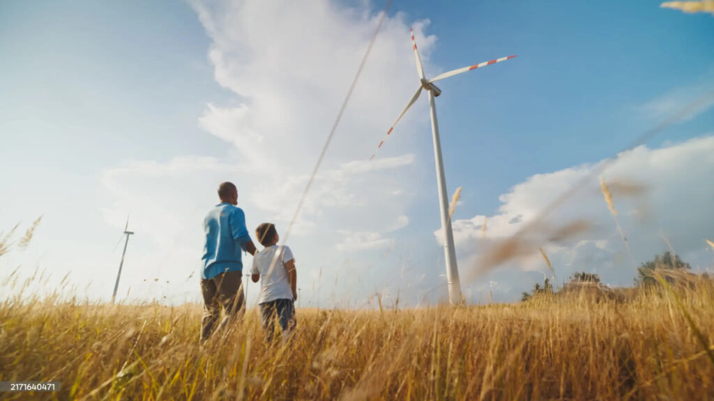 A man and a young boy stroll hand in hand through a golden wheat field, gazing at towering wind turbines under a clear, expansive sky.